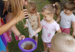 Pani tworzy magiczną banieczkę z dymkiem na rączce Aluni. Pozostałe dzieci z zaciekawieniem obserwują.