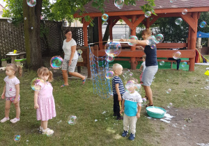 Ciocia Ola tworzy wiele ogromnych banieczek mydlanych dla maluszków. Dzieci z radością próbują je łapać.