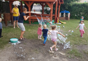 Ciocia Ola tworzy wiele ogromnych banieczek mydlanych dla maluszków. Dzieci z radością próbują je łapać.